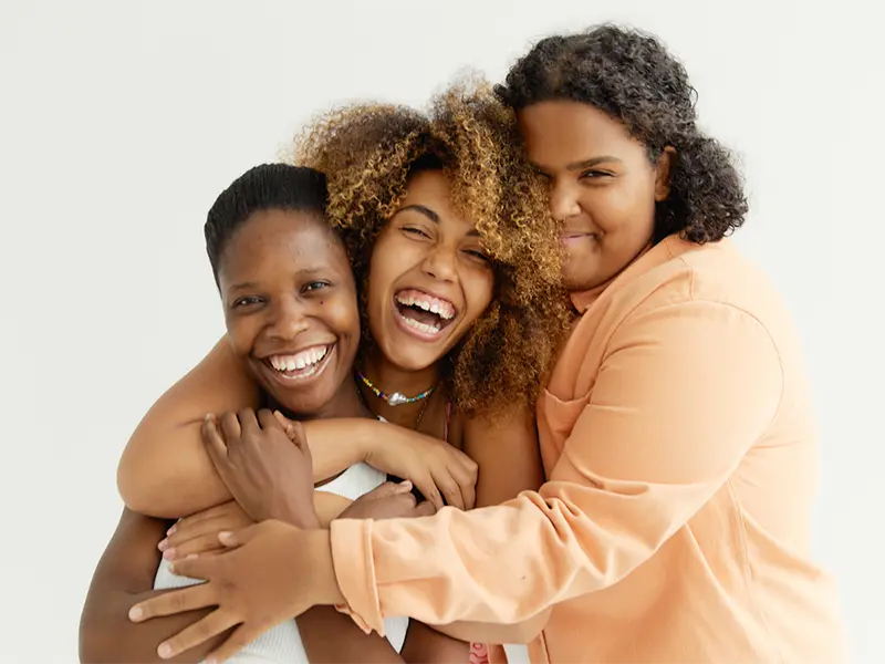 Three smiling individuals standing close together in a friendly embrace. The person on the left is wearing a white sleeveless top and a floral skirt, while the person in the middle has curly hair and a pink-patterned top. The person on the right has curly hair and is dressed in a peach-colored long-sleeve shirt. They are posing against a plain white background.